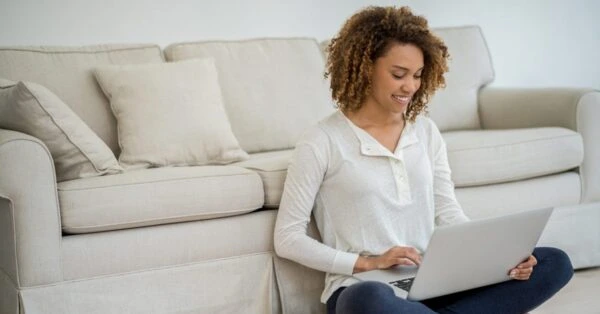 A woman sitting cross-legged on the floor in front of her couch while on her computer.